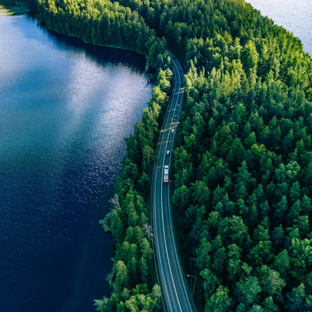 Aerial view of a truck driving on a highway surrounded by lush green forest and blue water