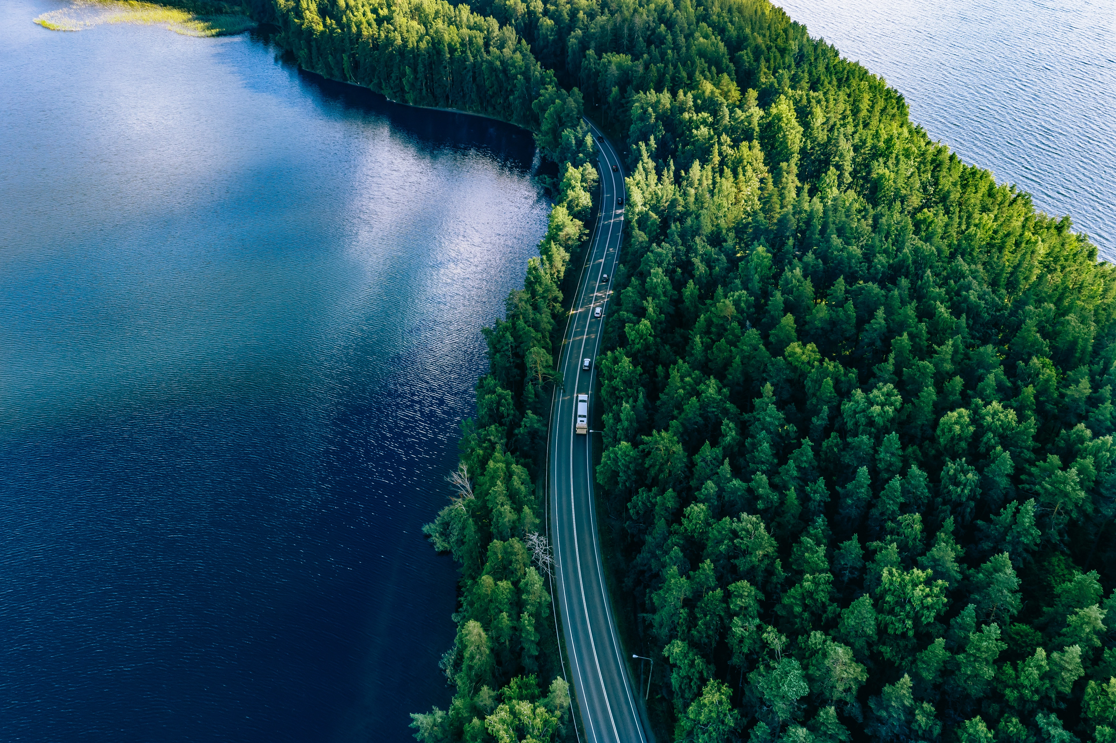 Aerial view of a truck driving on a highway surrounded by lush green forest and blue water