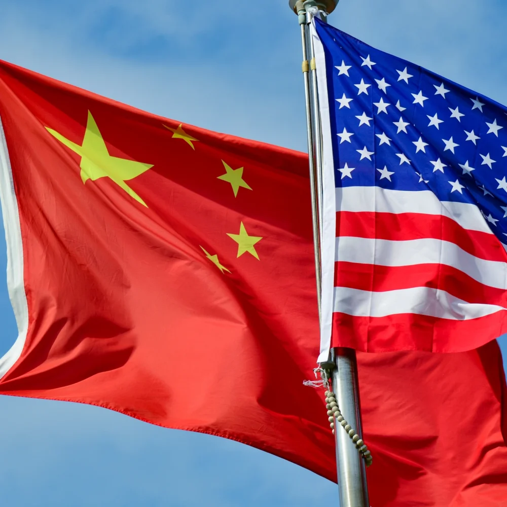 Chinese national flag and United States flag waving side by side on flagpoles against a clear blue sky, symbolizing international relations and diplomacy.