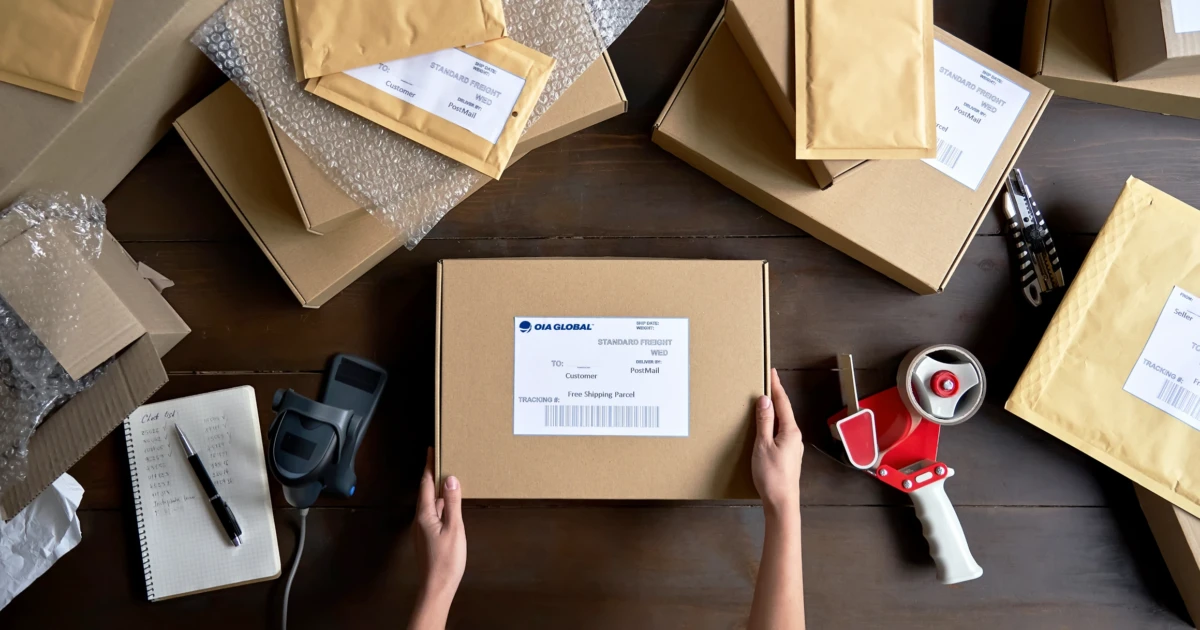 Hands preparing a cardboard shipping box with a printed label on a wooden table surrounded by bubble mailers, tape dispenser, and packing supplies.
