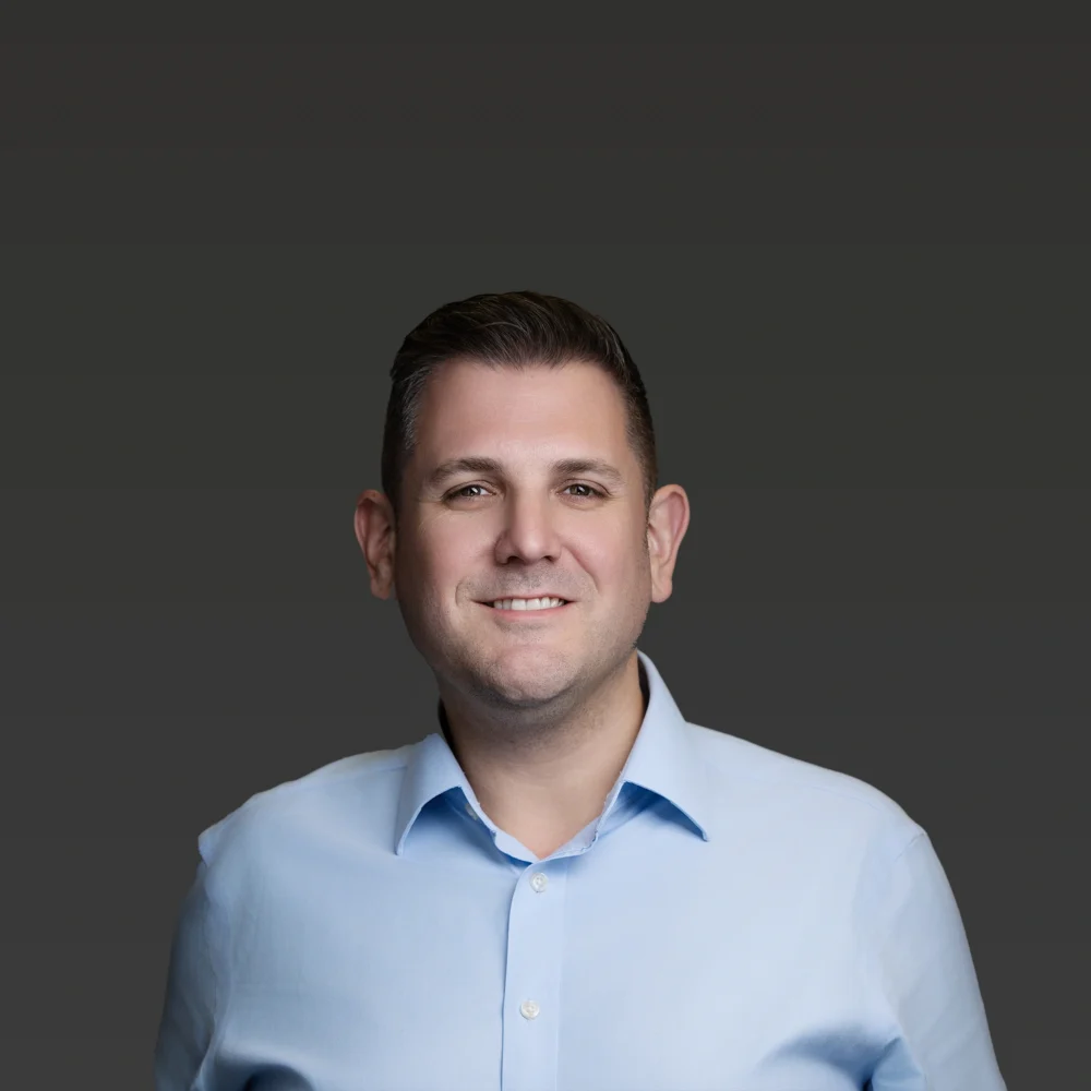 Professional corporate headshot of a business professional in a light blue shirt against a dark neutral background.