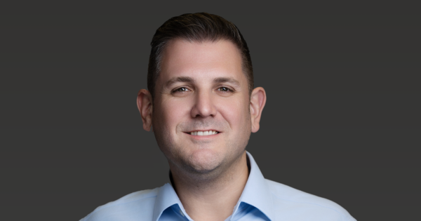 Professional corporate headshot of a business professional in a light blue shirt against a dark neutral background.