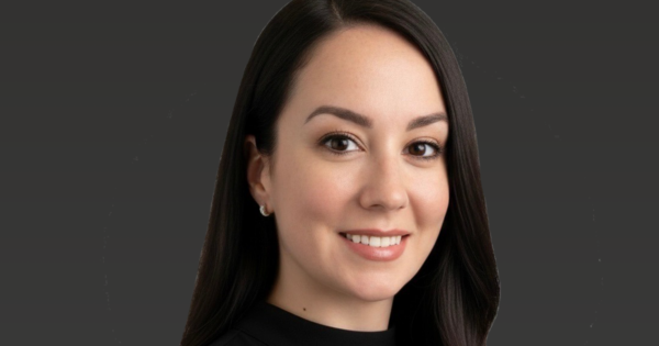 Professional corporate headshot of a business professional with long hair wearing a black top against a neutral gray background.