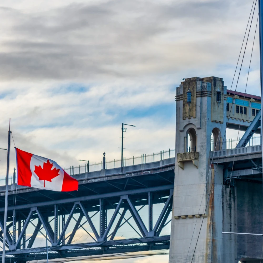 Canadian flag flying in front of a steel bridge structure with architectural towers and waterfront setting.